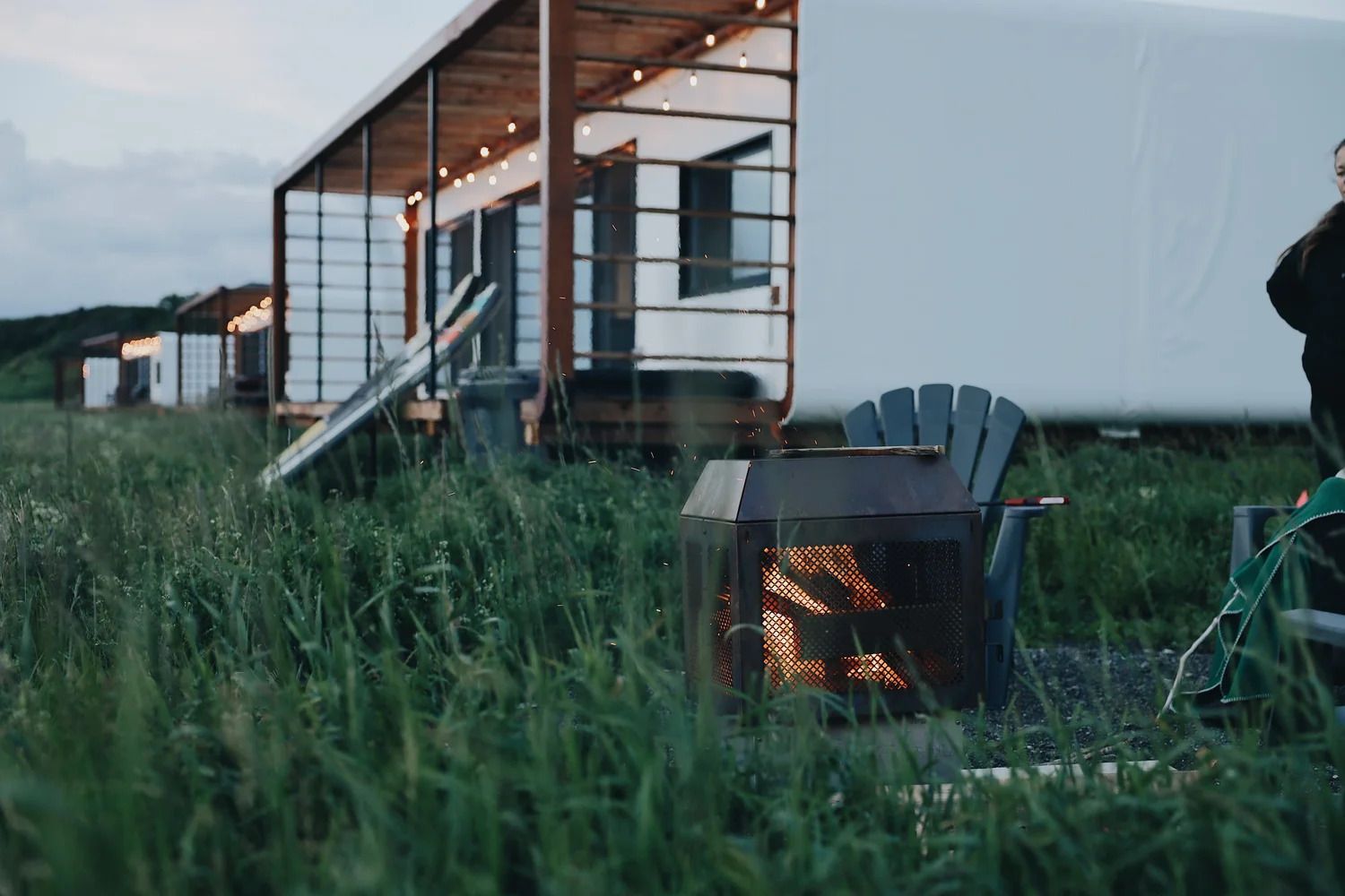 Veranda met een brandende vuurplaats en blauwe stoelen naast een huis, gezien door het hoge gras in de schemering.