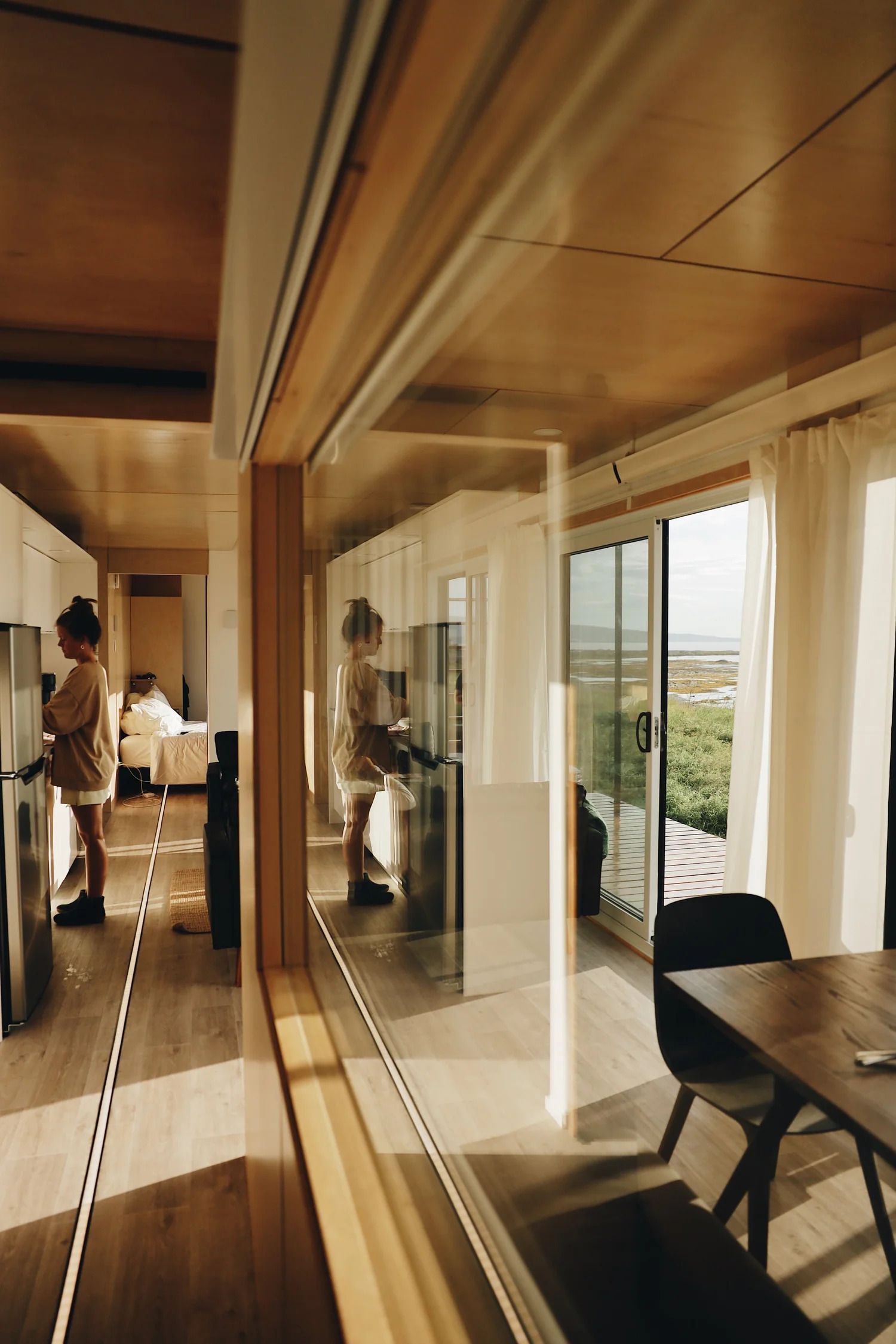Moderne appartementshal met spiegelwand, houten plafond, glazen deuren en een persoon die bij een eettafel staat.