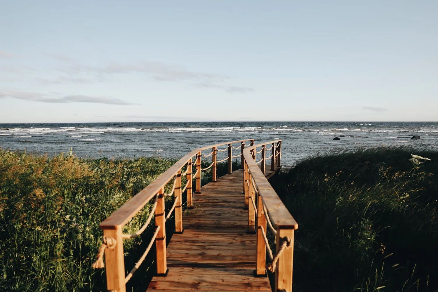 Een houten vlonderpad leidt door de duinen naar een winderige oceaan onder een bleke hemel.