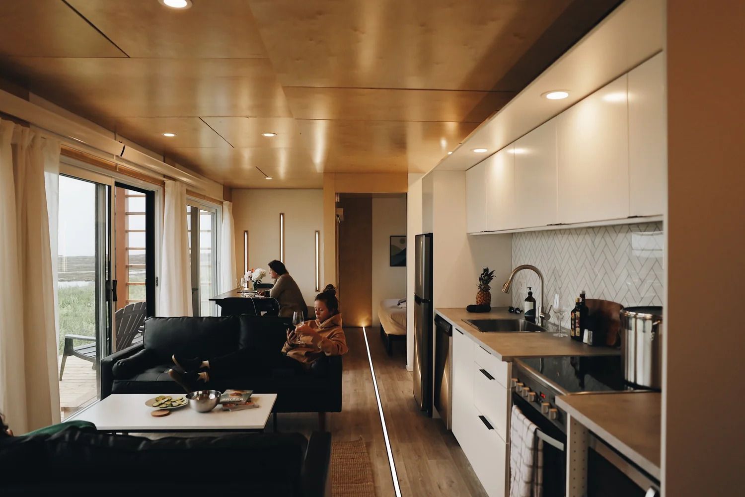 Moderne open woonkamer en keuken met zwarte banken, houten plafond en grote ramen.