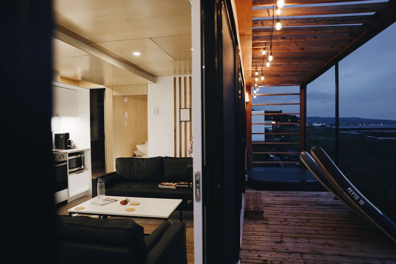 Modern appartementbalkon bij schemering, met de woonkamer zichtbaar door de openslaande deuren en uitzicht op de stad en de zee.