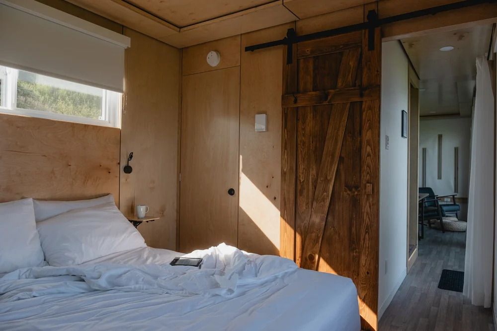 Zonnige slaapkamer met wit bed, houten wanden en kledingkast, en hal zichtbaar door openslaande deur.