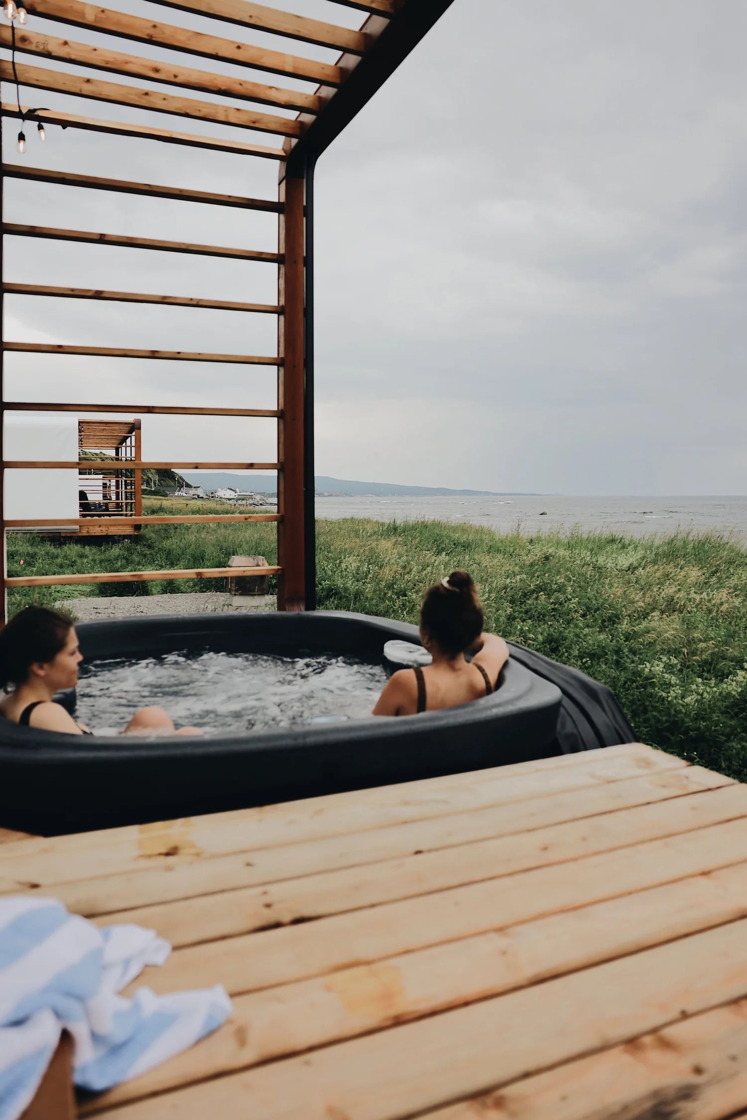 Twee mensen ontspannen bij Panora Loges Fluviales in een jacuzzi op een houten terras met uitzicht op de oceaan, onder een pergola.