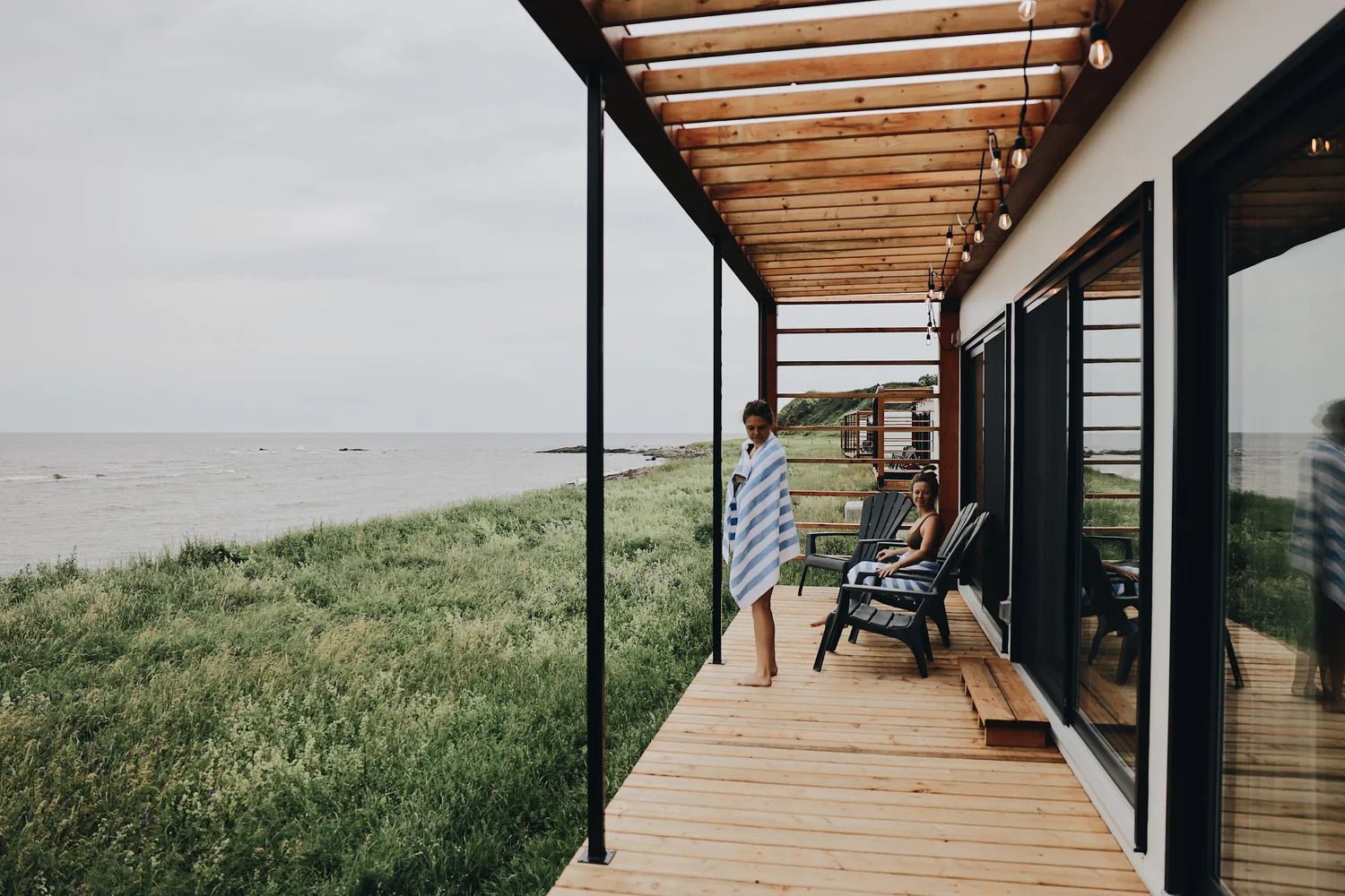Een persoon staat op een houten veranda naast een grasrijke oever met kalm water onder een overdekt terras.
