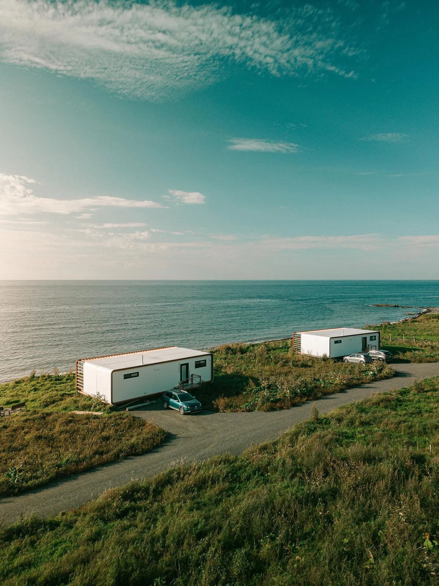 Twee witte caravans langs een met gras begroeide kustweg, met uitzicht op een kalme zee onder een blauwe hemel.