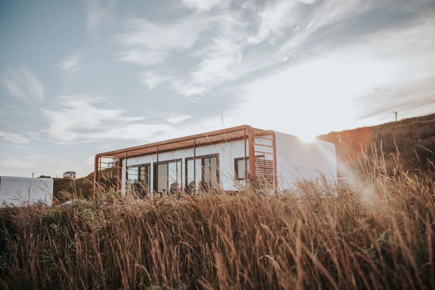 Moderne blokhut met grote ramen, gelegen naast hoog gras onder een bewolkte hemel.