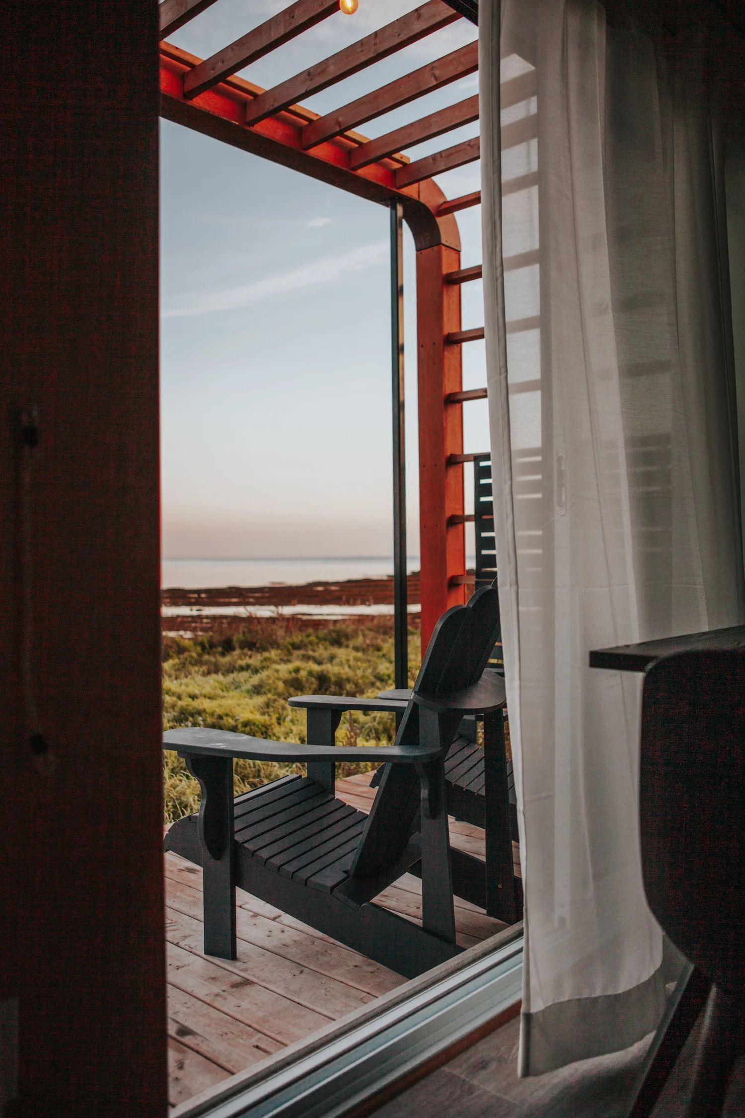 Uitzicht vanaf het balkon door een schuifdeur, met donkere stoelen, rode pergolabalken en een zonsondergang boven het water.