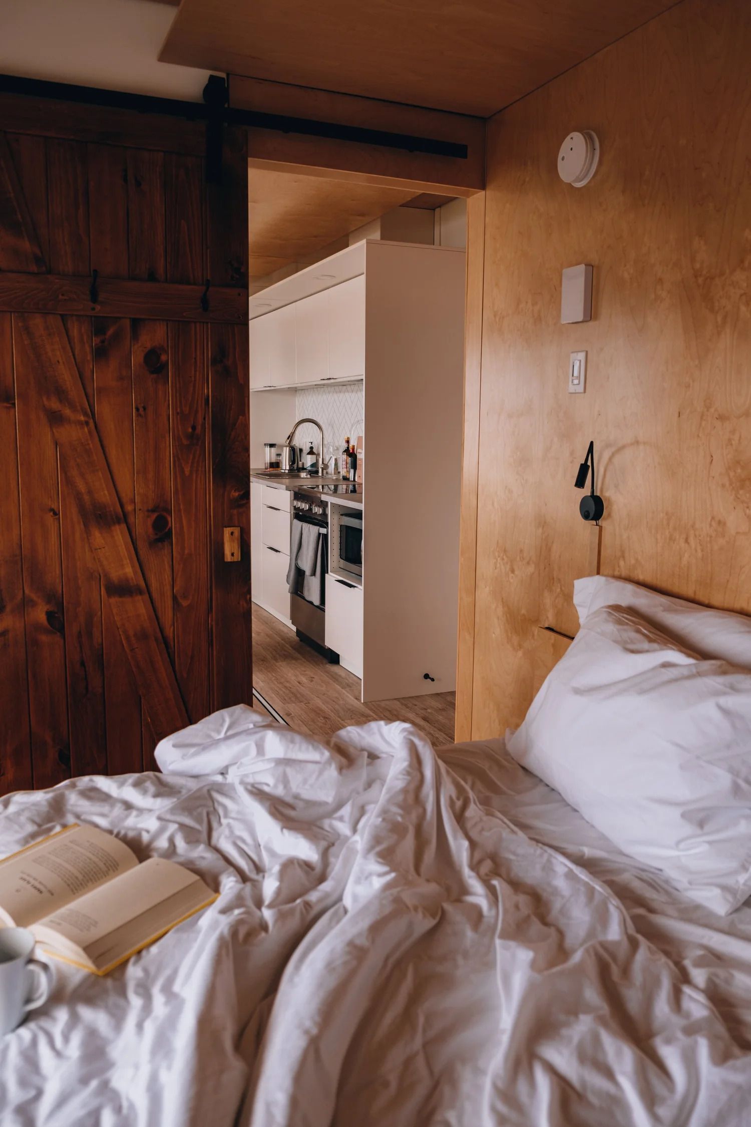 Een onopgemaakt wit bed in een slaapkamer met houten lambrisering, met uitzicht door een open deur op de keuken.