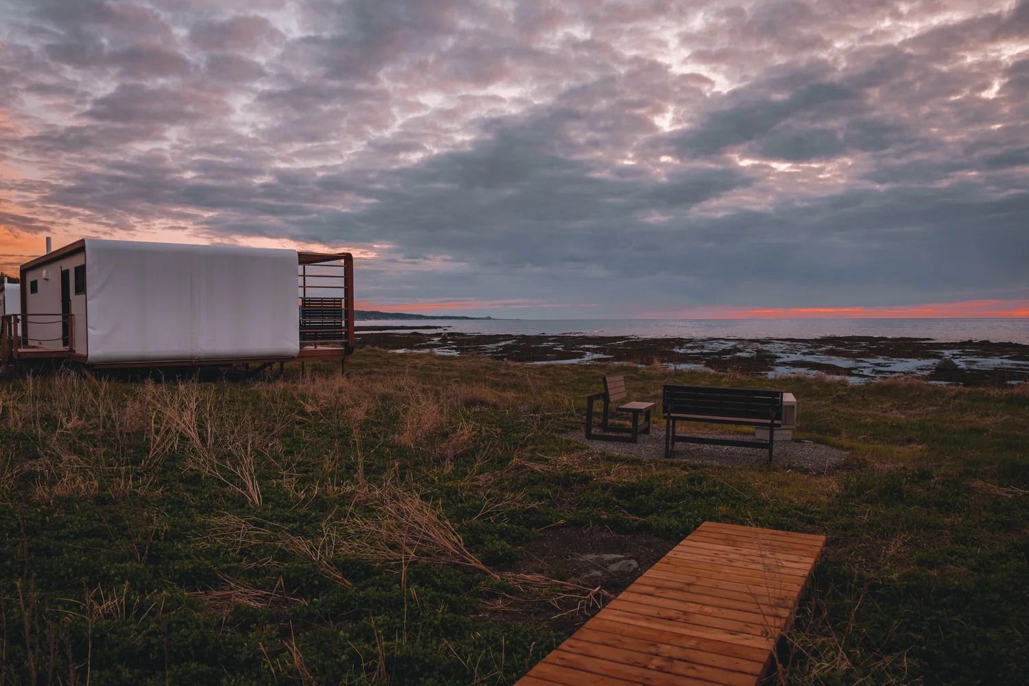 Een houten wandelpad langs de kust bij zonsondergang met een bankje, grasduinen en een klein wit hutje onder een bewolkte hemel.