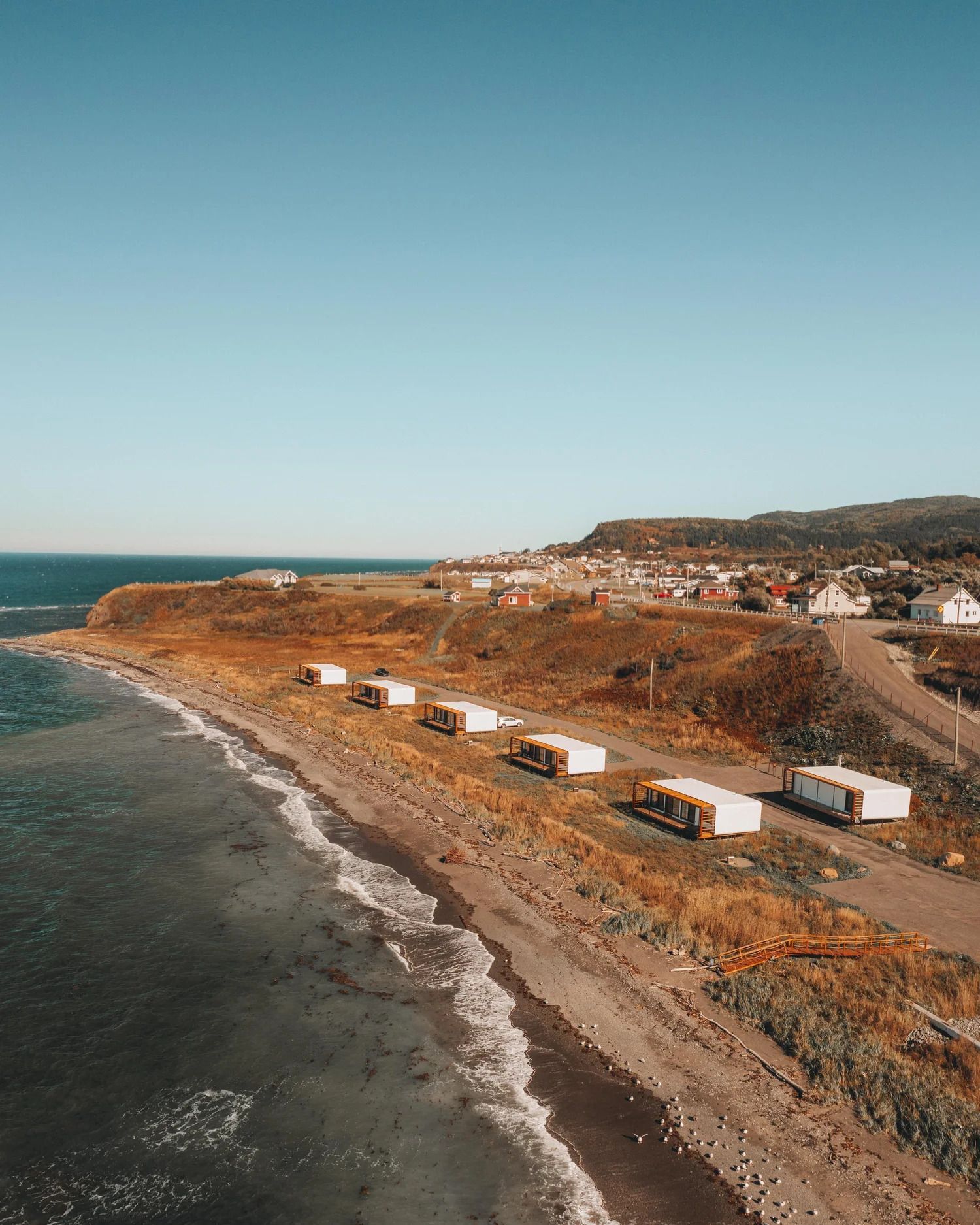 Luchtfoto van een rotsachtige kustlijn met oceaangolven, caravans op een weg en huizen op een heuvel.