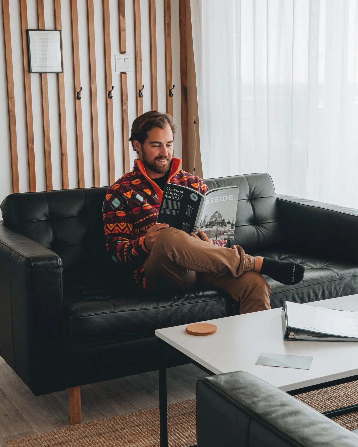 Een man zit op een zwarte bank en leest een tijdschrift in een lichte woonkamer.