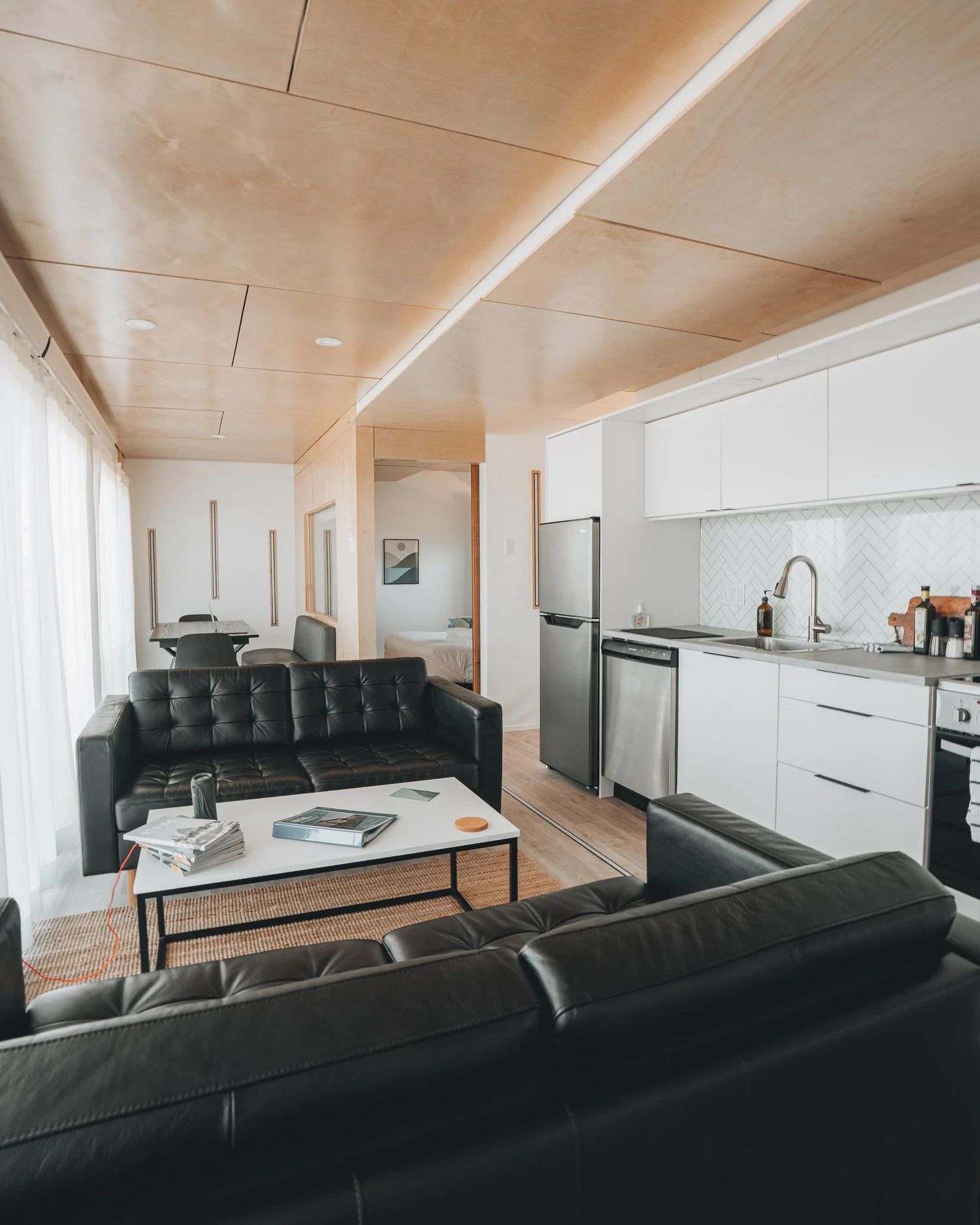 Moderne open woonkamer en keuken met zwarte banken, witte kasten en een houten plafond.