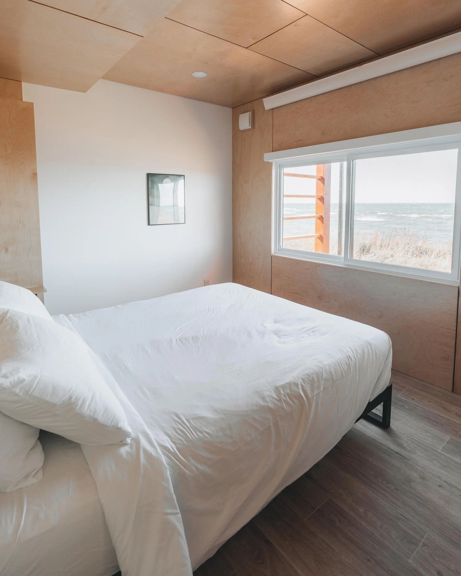 Minimalistische slaapkamer met wit bed, houten plafond en groot raam met uitzicht op het landschap.