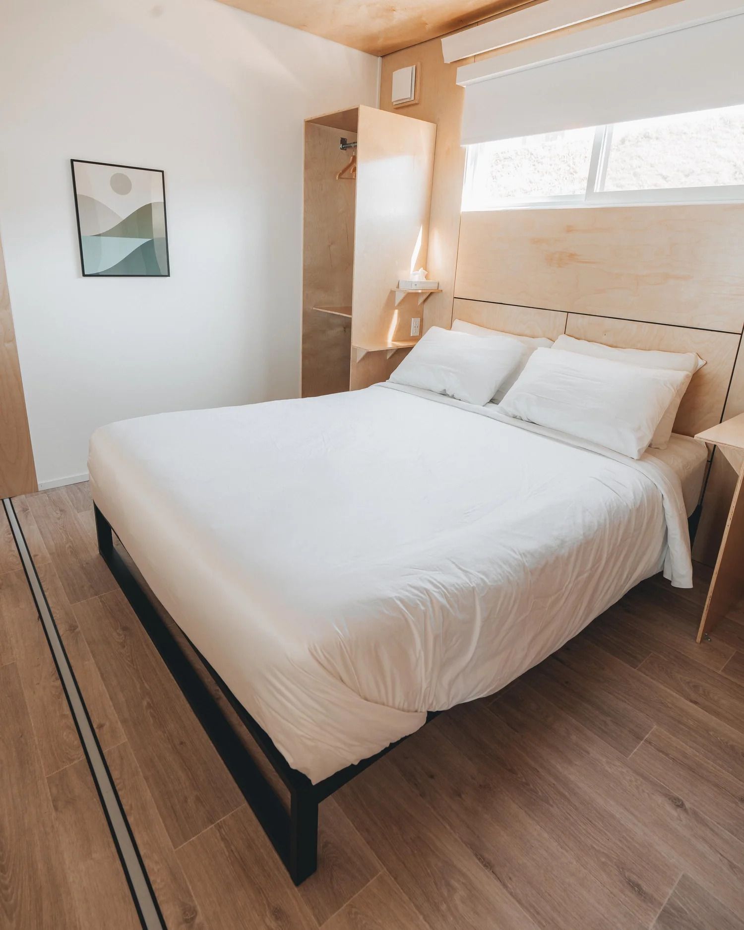 Lichte slaapkamer met een keurig opgemaakt wit bed, houten vloer en een raam boven het hoofdeinde.