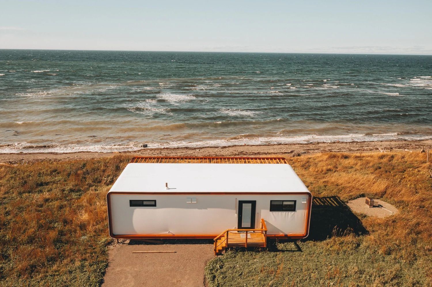 Een kleine, witte hut aan de kust met oranje stoelen, uitkijkend op de oceaan en een grasrijk strand.