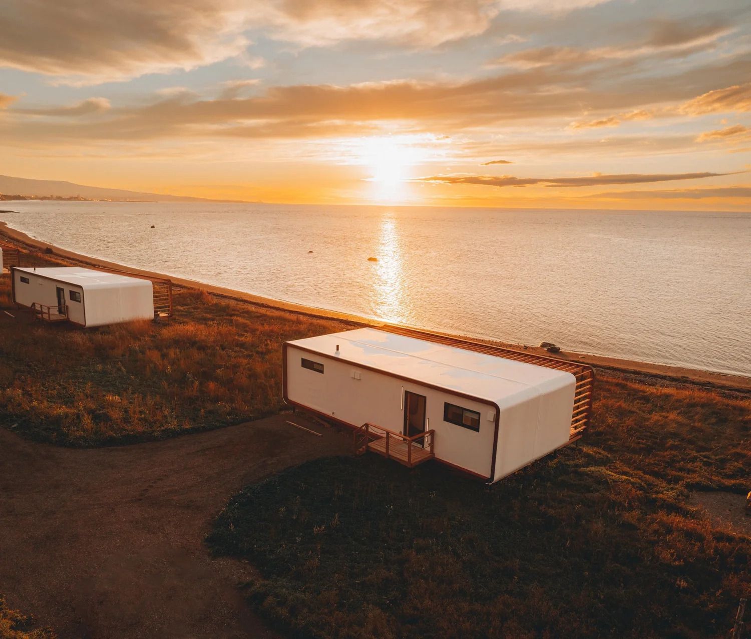 Twee witte caravans op een klif aan de kust bij zonsondergang, met uitzicht op kalm water en een oranje hemel.