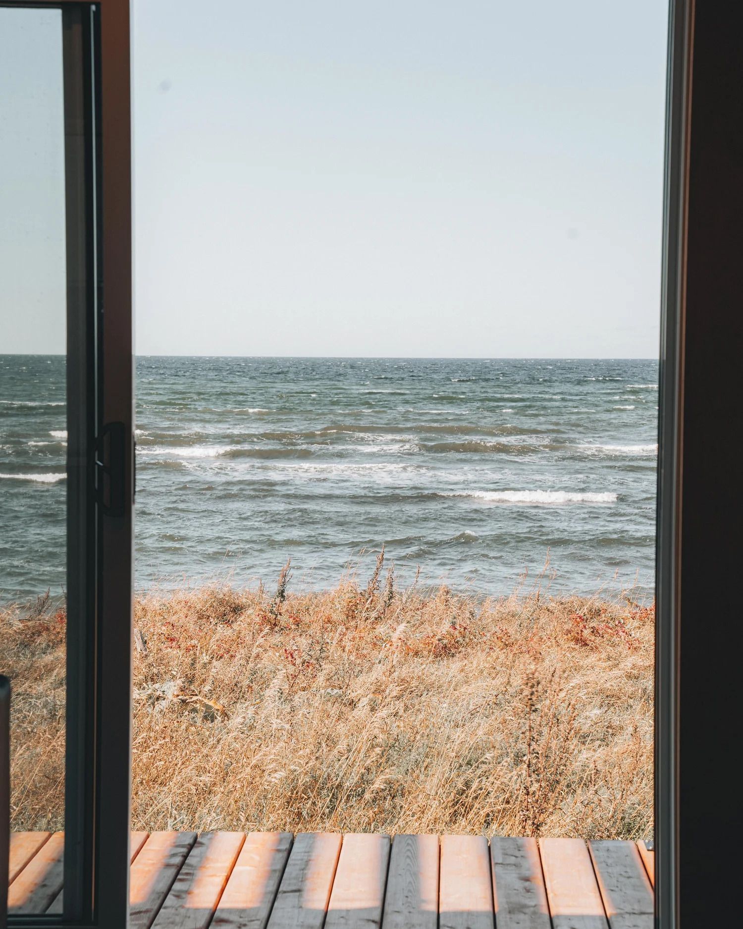 Uitzicht door een raam op een strand met droog gras, golven en een houten vlonder op de voorgrond.