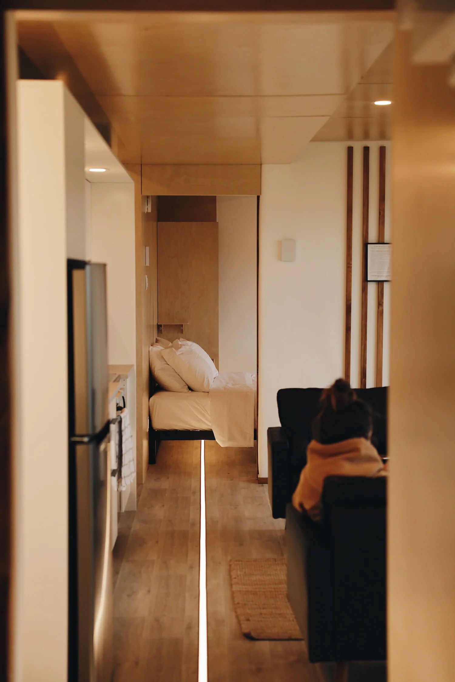 Smalle houten gang die leidt naar een bed, met een bank en warme verlichting in een moderne kamer.