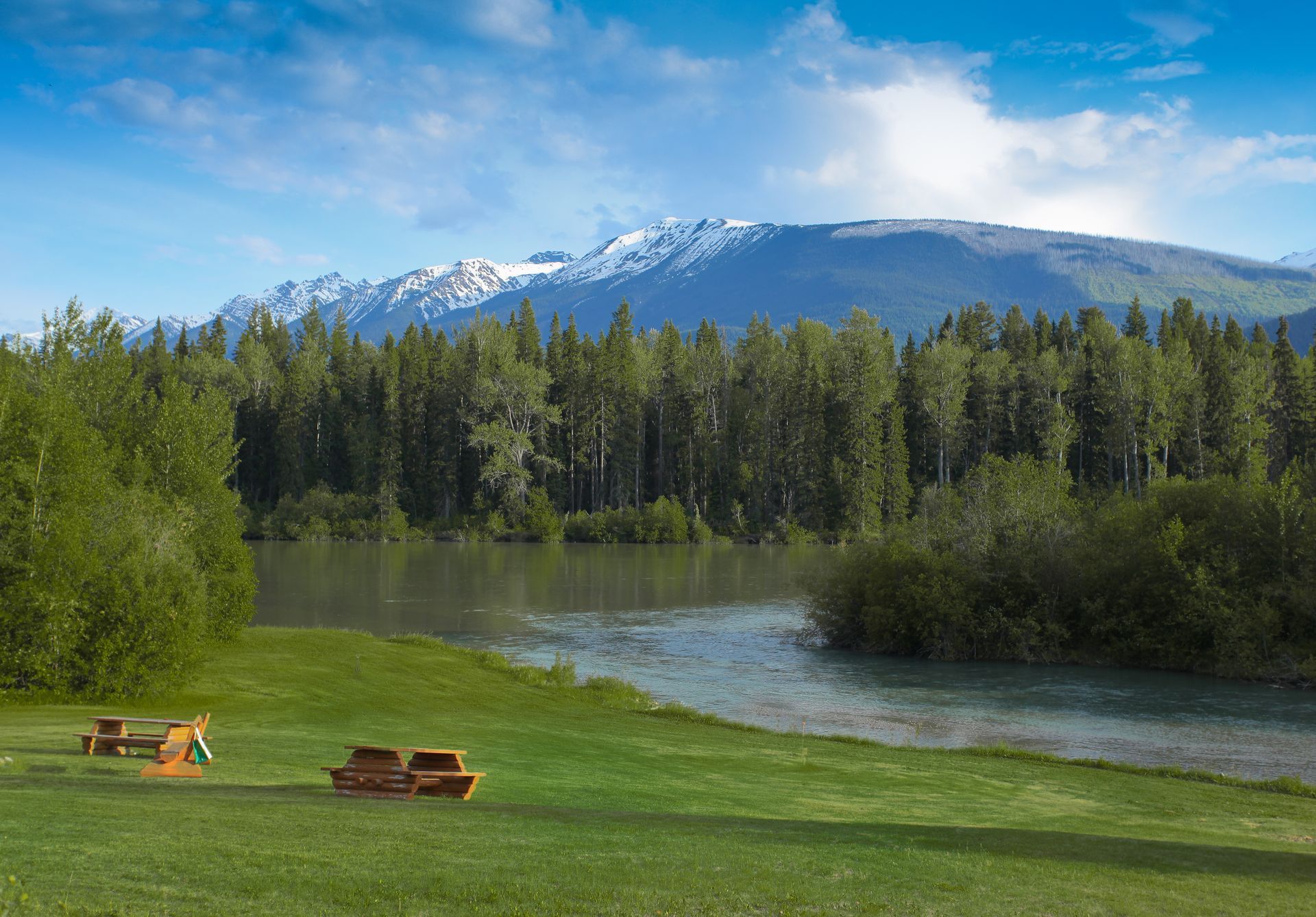 Grasrijke oever aan de North Thompson River nabij Valemount met picknicktafels, kalm water, naaldbomen en besneeuwde bergen onder een blauwe hemel.