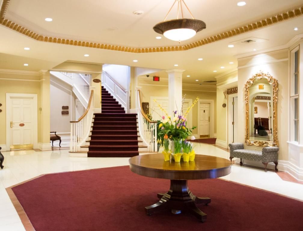 Elegante hotellobby met een imposante trap, ronde tafel, bordeauxrood tapijt en kroonluchterverlichting.