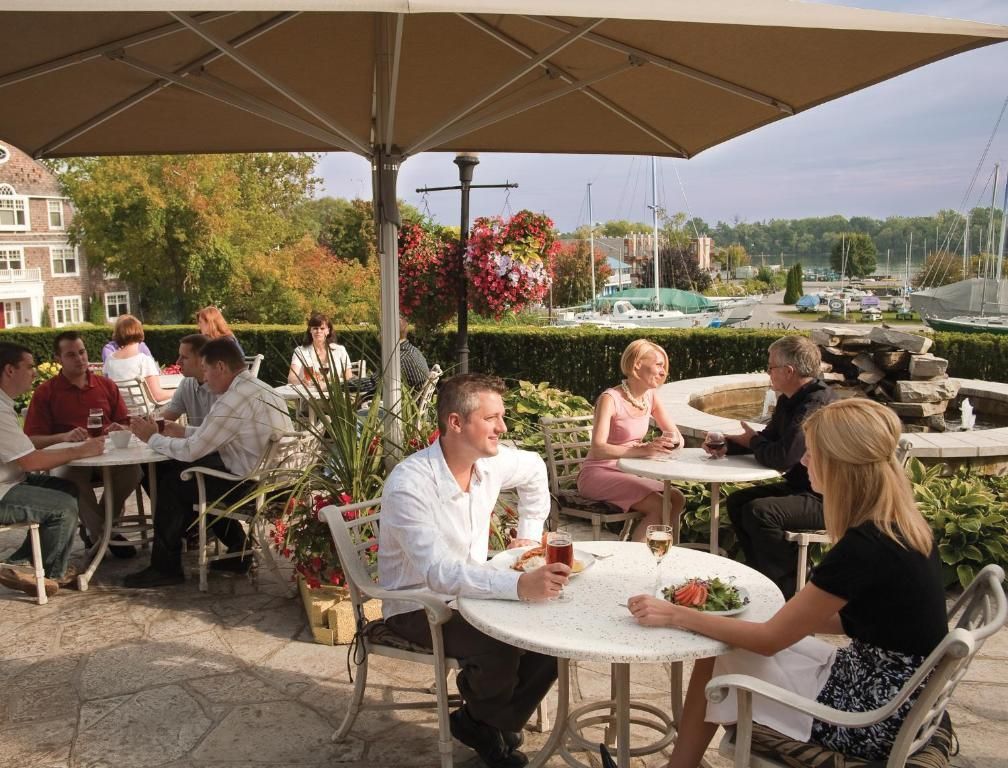 Mensen dineren op een terras van een café met witte tafels, parasols en uitzicht op de jachthaven.