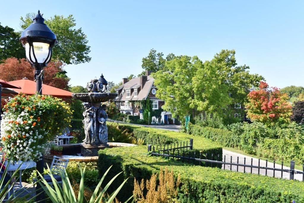 Een formele tuin met strak gesnoeide hagen, een fontein, een lantaarnpaal en een huis op de achtergrond onder een helderblauwe hemel.