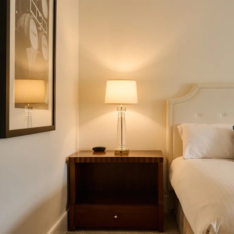Gezellige slaapkamerhoek met lamp op houten nachtkastje, wit bed en ingelijste muurdecoratie.