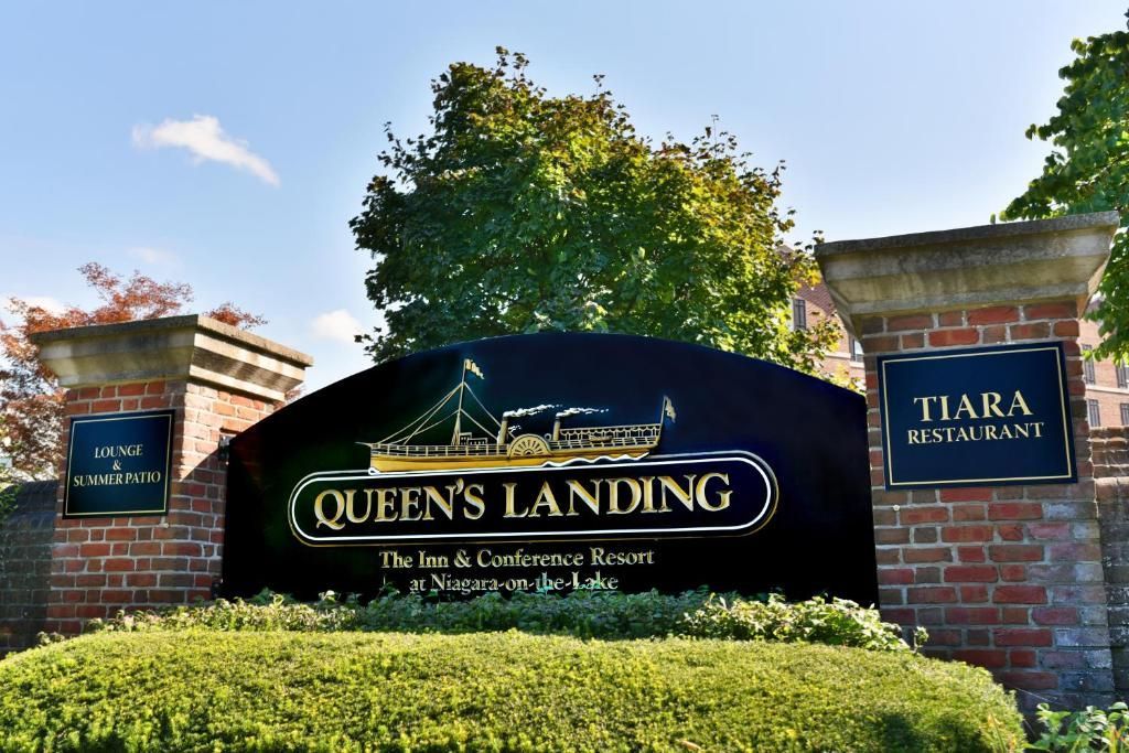 Het entreebord van Queens Landing met bakstenen pilaren en groene beplanting onder een blauwe hemel.