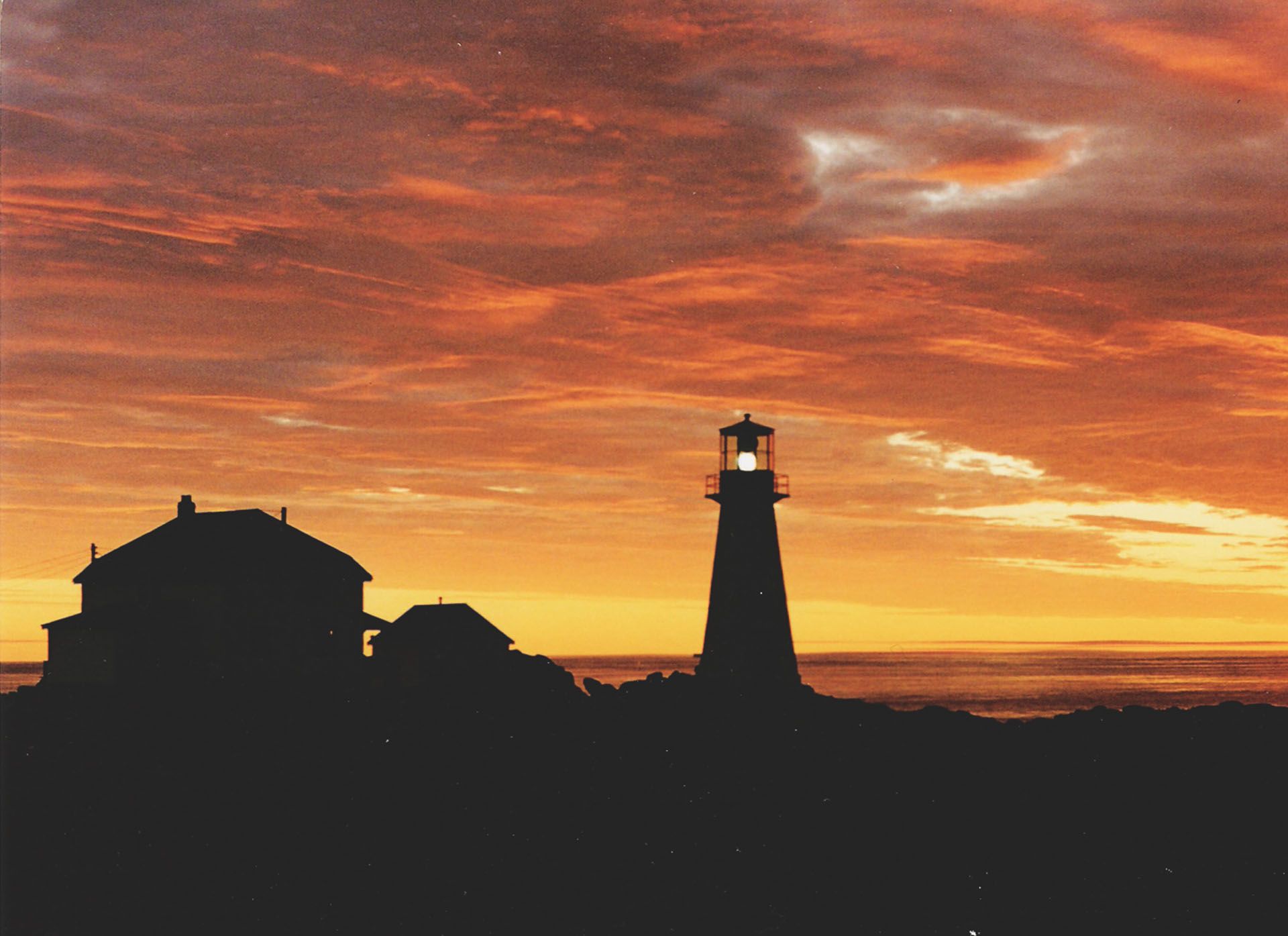 Silhouet van een vuurtoren en gebouwen bij zonsondergang onder een dramatische oranje hemel.