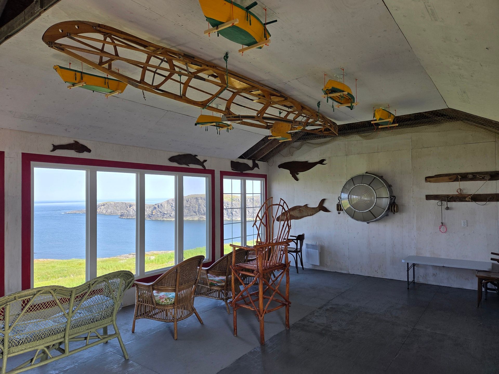 Binnenkamer van Quirpon Lighthouse Inn met ramen die uitzicht bieden op de oceaan, rieten stoelen en decoraties met vissen en boten aan de muren en het plafond.