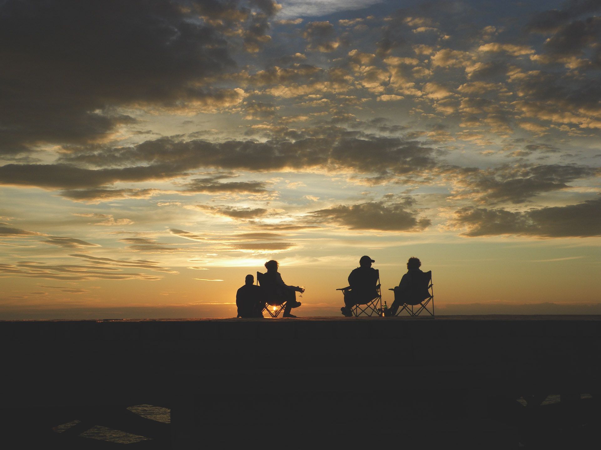 Silhouetten van drie mensen die op stoelen zitten en naar een zonsondergang boven een vlakke horizon kijken.