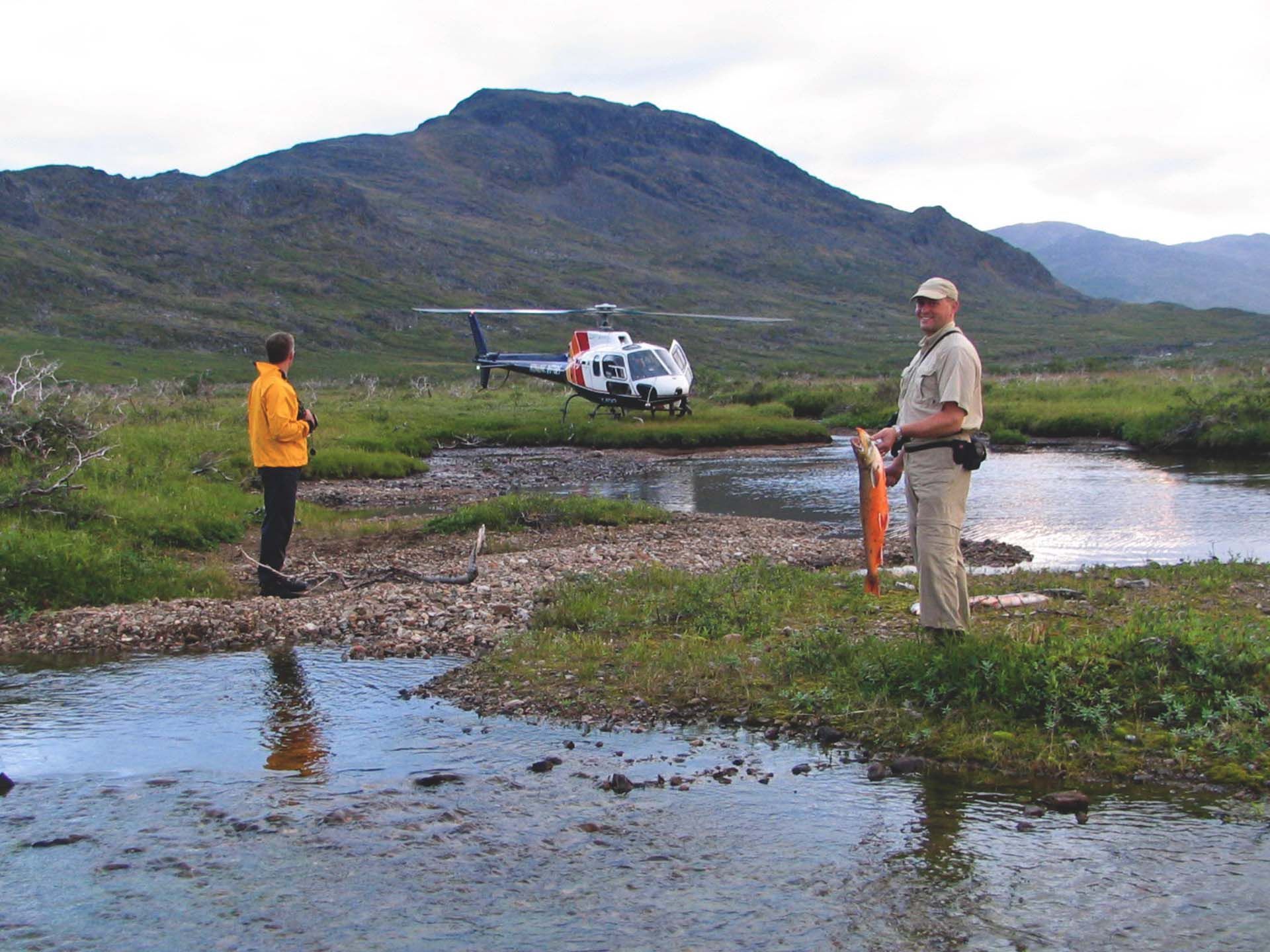 Helikopter landt vlakbij twee mensen in een modderig moerasgebied met bergen op de achtergrond.