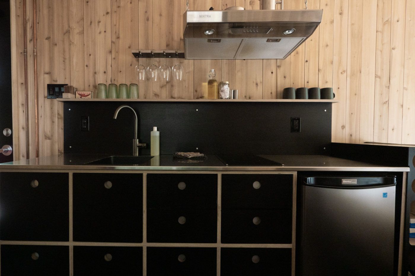 Moderne keuken met houten wanden, zwarte kasten, roestvrijstalen afzuigkap, spoelbak en compacte koelkast.