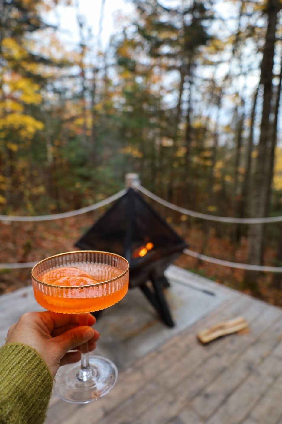 Hand die een sinaasappelsap vasthoudt bij een vuurplaats op een houten terras in het bos.