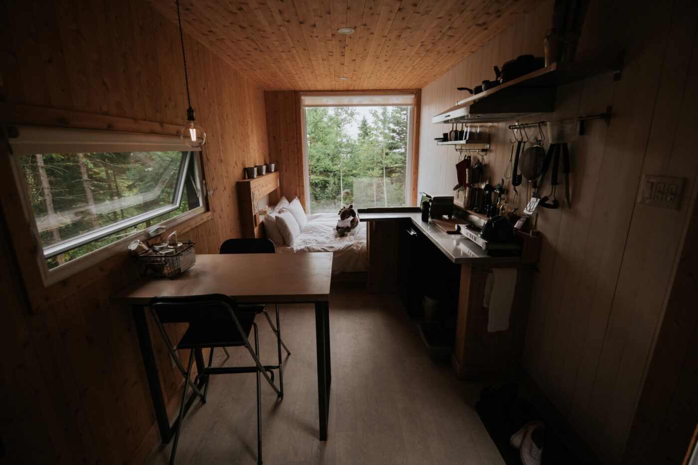 Gezellig interieur van een kleine houten hut met tafel, planken, kitchenette en een raam met uitzicht op het groen.