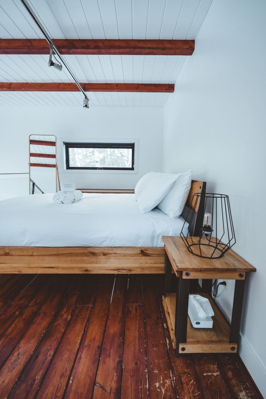 Kleine, rustieke slaapkamer met wit bed, houten vloer, zichtbare plafondbalken en een nachtkastje.