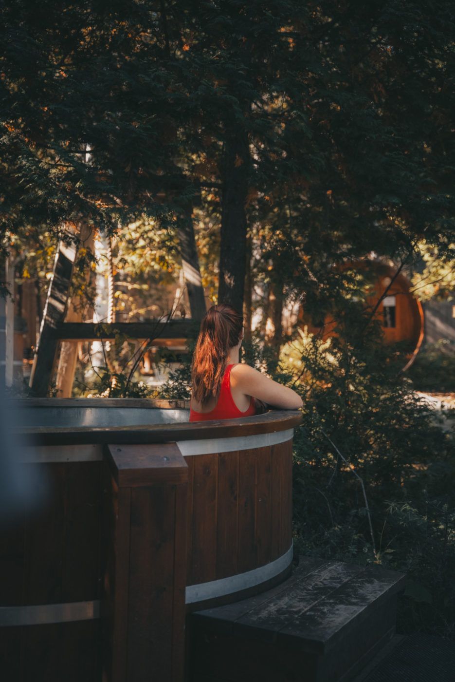 Vrouw in een rode jurk leunend tegen een houten jacuzzi in een groene, buiten gelegen spa van Repere Boreal in Canada.