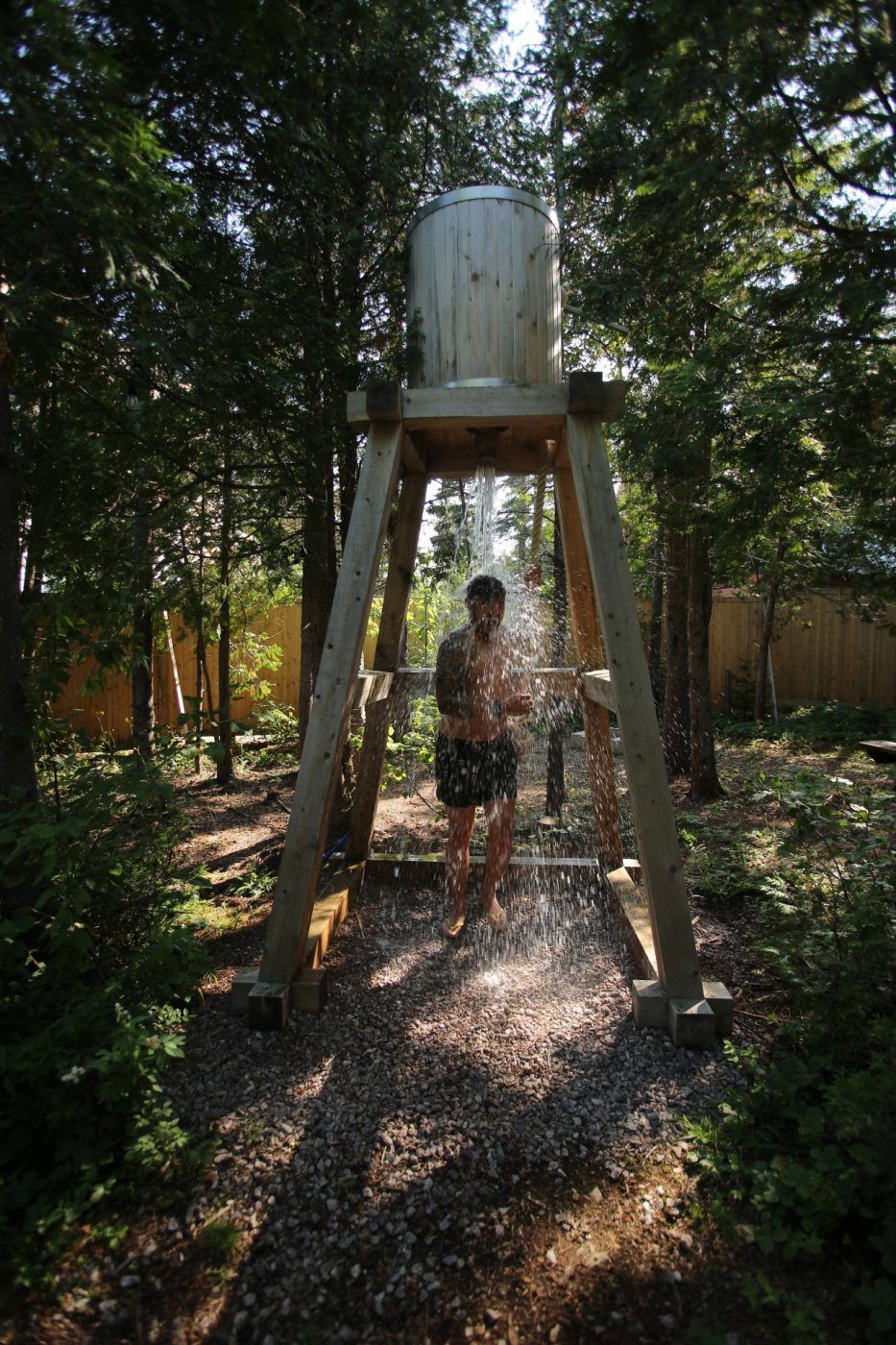 Een persoon staat onder een watertank op een schaduwrijk tuinpad.