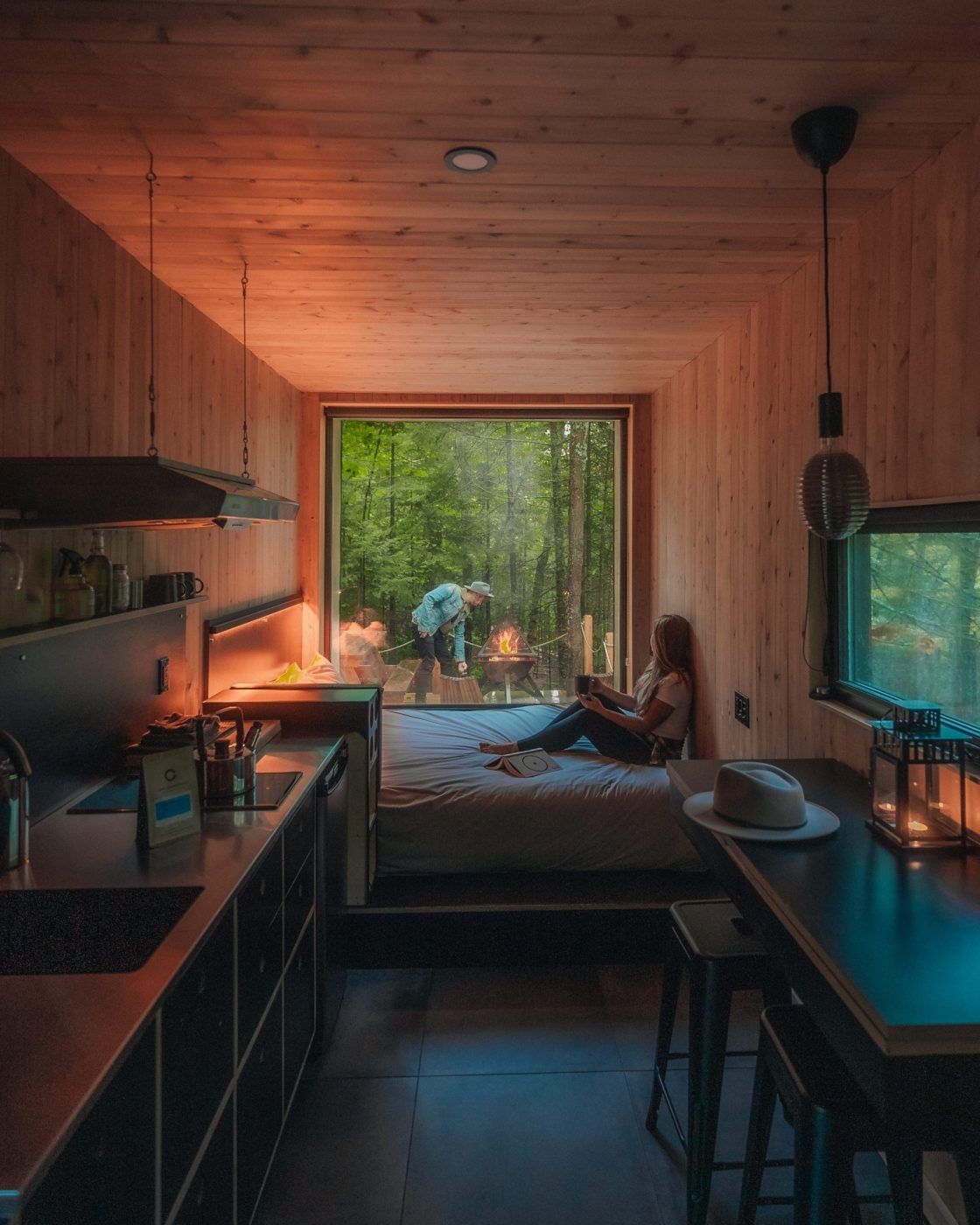 Gezellige, met hout beklede keuken in een blokhut, met iemand die ontspannen bij een raam zit met uitzicht op een groen bos.