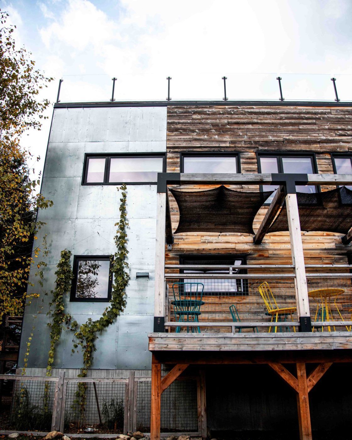 Modern gebouw met metalen en houten gevelbekleding, klimop op de muur en een overdekt balkon onder een heldere hemel.