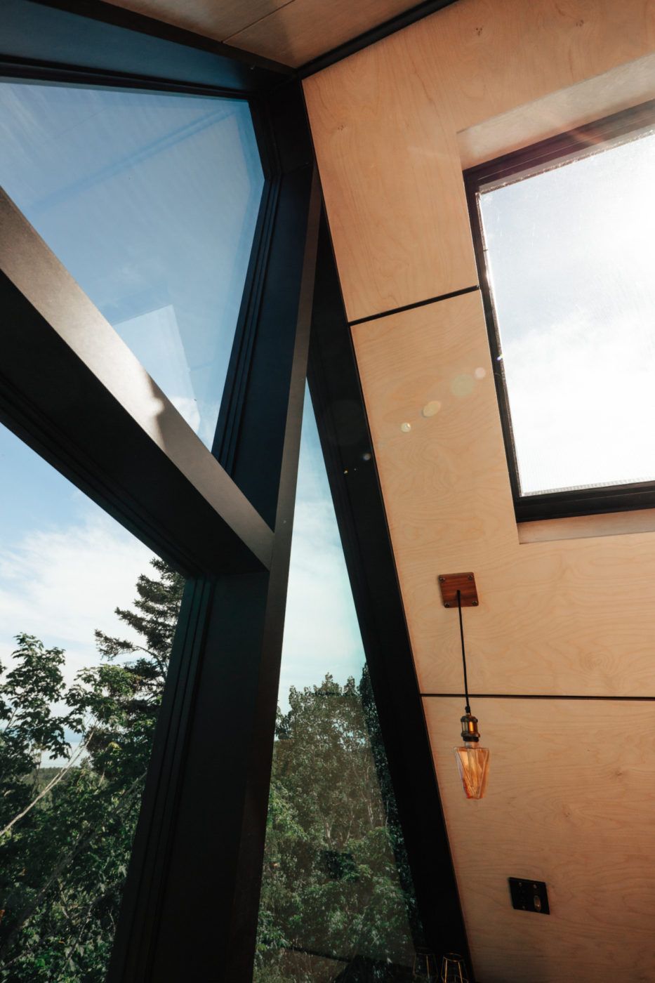 Modern interieur met schuine glazen ramen en beige muren, met uitzicht op bomen buiten en een hangende gloeilamp.