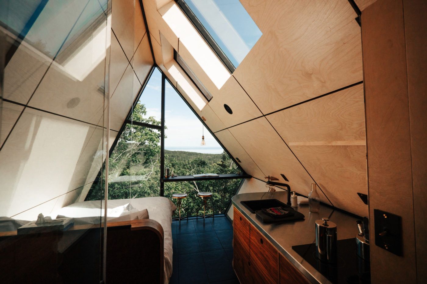 Klein, modern interieur van een blokhut met dakramen, houten lambrisering, een bed en uitzicht op bomen door een groot raam.