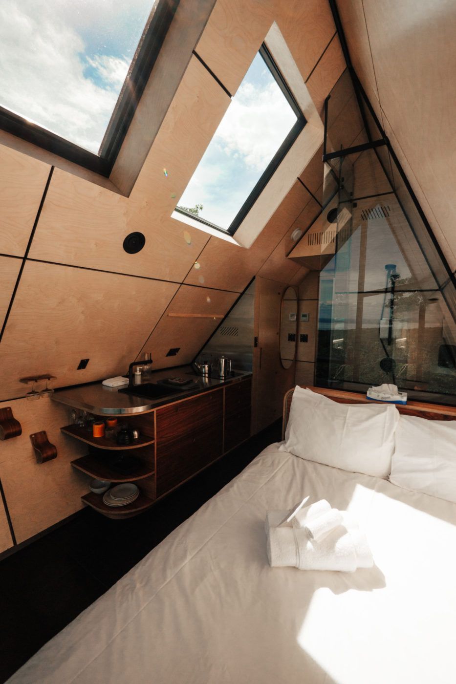Kleine loftslaapkamer met dakramen, wit bed en compacte kitchenette naast een glazen scheidingswand.