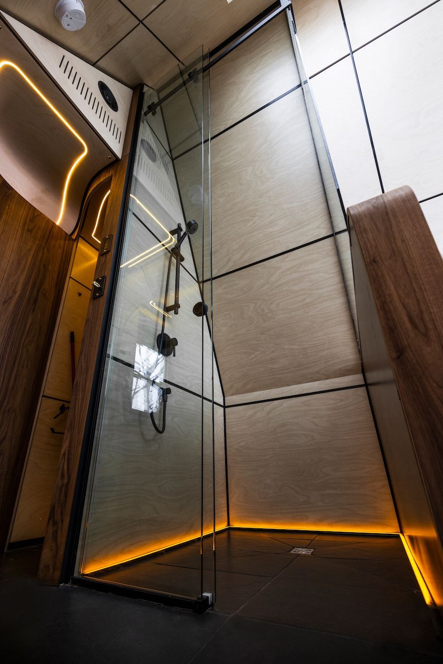 Moderne douchecabine met glazen deur, beige betegelde wanden en warme oranje vloerverlichting.