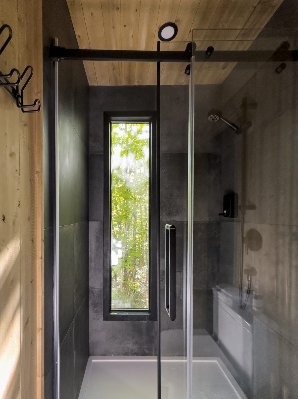 Moderne douchecabine met glazen deur, donkere wanden en een smal raam dat natuurlijk licht binnenlaat.
