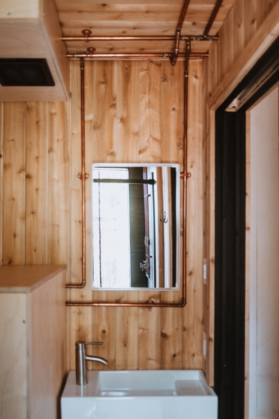 Kleine badkamer met houten lambrisering, een spiegel, wastafel en zichtbare koperen leidingen.