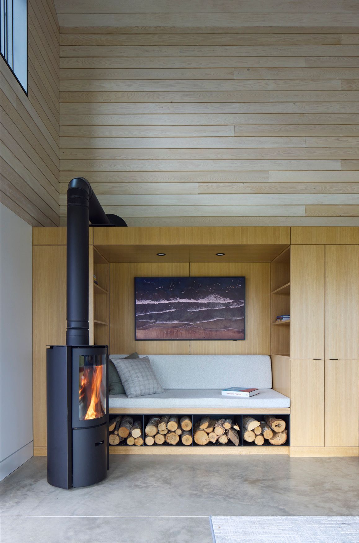 Minimalistische, met hout beklede nis met een ingebouwde bank, opgestapeld brandhout en een zwarte houtkachel bij een raam.