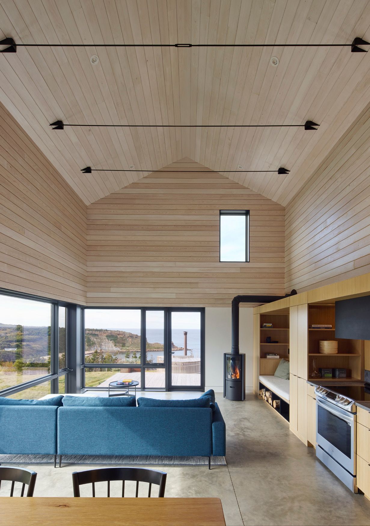 Moderne woonkamer met blauwe bank, houten plafond, grote ramen en open keuken.