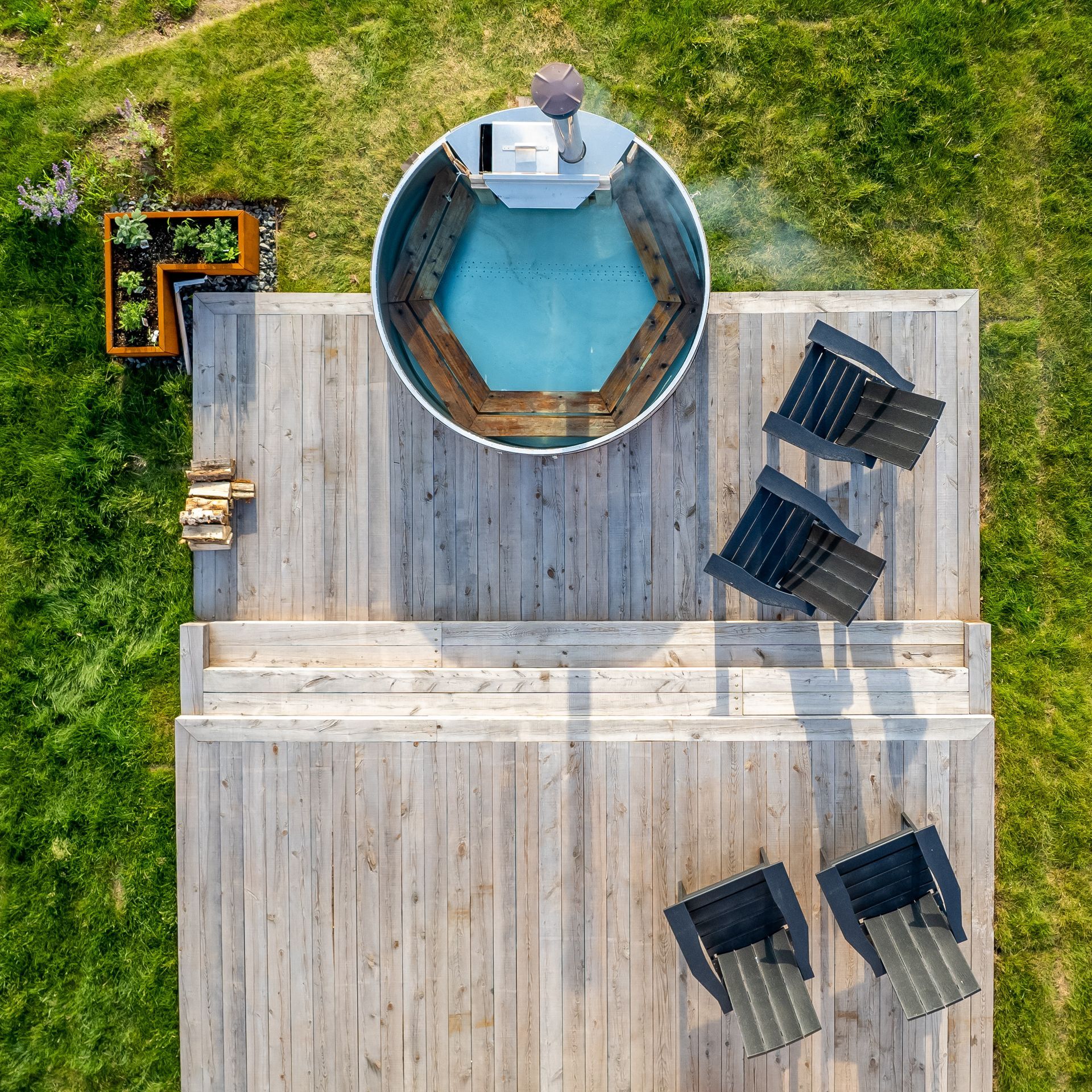 Bovenaanzicht van een houten terras met een ronde blauwe jacuzzi en ligstoelen op het gras.