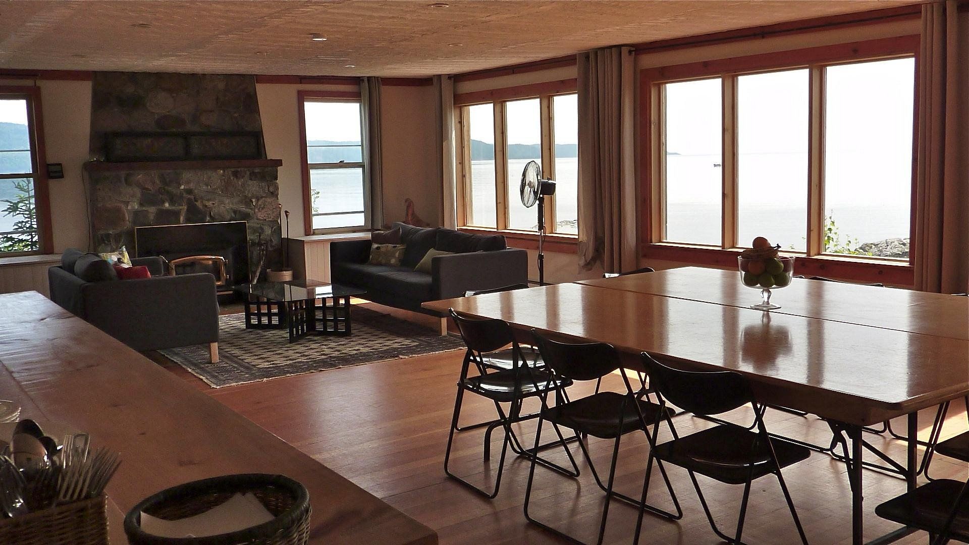 Moderne gezamenlijke ruimte van Rock Island Lodge met grote ramen, stenen open haard en een lange houten tafel met uitzicht op de oceaan.